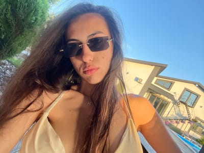 The image features a young woman taking a selfie outdoors. She has long, dark brown hair, is wearing sunglasses, and a light-colored top. Her face is the primary focus, and she appears to be looking towards the camera. She has some minor blemishes on her skin.
The background is a bright blue sky, suggesting it is daytime and the weather is clear. To the left of the frame, part of a building is visible, with windows and a ladder leading up to a balcony. There are also some potted flowers on the balcony. To the right, there is a lush green hedge.
The orientation of the image is rotated, making it appear as if the subject is lying down or the camera is held upside down. This perspective, combined with the outdoor setting, suggests a relaxed or casual moment, possibly by a pool or in a garden. No text is visible in the image. The location context provided is Novooleksandrivka, Ukraine.