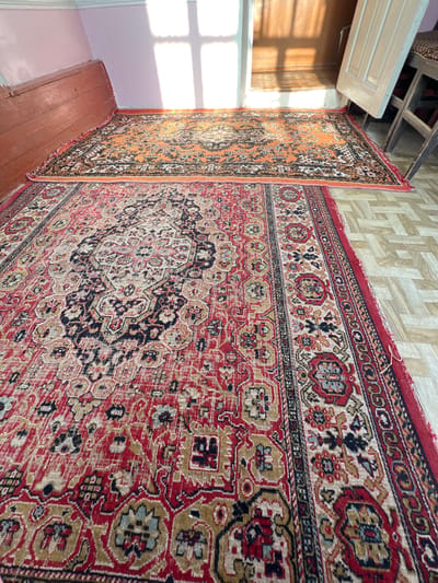 The image shows two Oriental-style rugs on the floor of a room. The rug in the foreground is predominantly red with intricate patterns in black, gold, and blue. The rug in the background is orange with a black and white pattern. Sunlight streams through a window, casting shadows on the floor and the rugs. A wooden door is ajar in the background, revealing a glimpse of another room. A portion of a wooden chair is also visible. The room appears to be sparsely furnished and may be in an older building. This scene is consistent with a domestic interior in Hazorasp, Uzbekistan, a region known for its traditional rug-making.