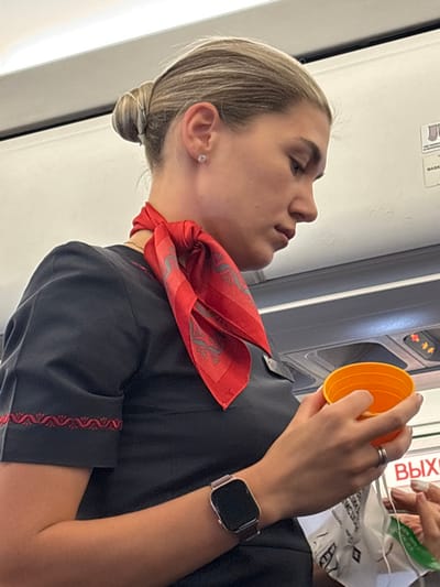 The scene depicts a female individual within an aircraft cabin, contextually located in or operating near Novogorelovo, Russia. The subject is a light-haired female, approximately 20-35 years old, with her hair neatly styled in a bun. She wears a dark, short-sleeved uniform top featuring a red patterned trim on the cuff of her left sleeve, and a red scarf with a repeating grey patterned motif tied around her neck. A dark nametag or badge is clipped to her uniform below the scarf. A small stud earring is visible in her right ear. On her left wrist, she wears a smartwatch with a rose gold-colored rectangular case and a dark mesh band. A simple silver-toned band ring is present on her left ring finger. She is holding an empty or partially filled orange plastic cup in her left hand, while her partially visible right hand appears to be manipulating a white plastic bag or wrapper. Her gaze is directed downwards and to her right. The background consists of the typical white overhead panels and storage compartments of an airplane interior. An overhead passenger service unit with air vents, call buttons, and an illuminated "no smoking" indicator is visible. A white sign with red Cyrillic text "ВЫХОД" (Exit) is visible on a bulkhead or door frame to the right.