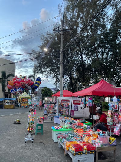 The image shows a street scene in Kuala Pidie, Indonesia. The shot is taken at ground level, facing a vendor stall along a street. The stall is covered with a red canopy and displays toys and other merchandise. A person in a red shirt appears to be attending the stall. In the foreground, bunches of colorful character balloons are for sale along with other colorful toys and goods displayed on carts and tables. A utility pole with numerous electrical wires is visible in the background. The sky is overcast with diffused sunlight. There is a building partially visible on the left.