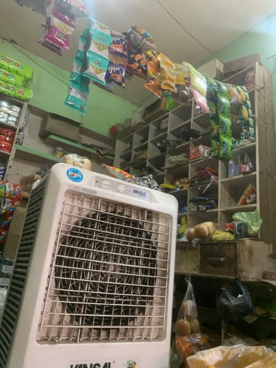 The image shows a small shop in Kanpur, India, located at the coordinates 26.44 latitude and 80.344 longitude. There is a white, metal, Vinsal brand air cooler in the center of the image. It is turned on. The air cooler is in front of a large wooden shelf unit. The shelf unit is filled with various items. Some of the items include: packaged snack foods, plastic bags, and other miscellaneous items. There is also a wooden drawer below the shelf unit with some loose items in it. There is a small plastic bag full of bread rolls on the shelf unit near the air cooler. On the wall above the shelf unit are rows of hanging snack food bags, which appear to be filled with different types of nuts, snacks, and spices. The wall is painted a light green color. The ceiling is white and appears to be textured.