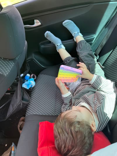 The image depicts a young child lying on their back in the backseat of a car. The child is wearing a striped long-sleeved shirt and grey pants with matching grey socks. Their legs are propped up against the car door, and they are holding a colorful pop-it toy. In the background, car seats and a black organizer with bottles and wrappers are visible. The child's head rests on a red pillow. The setting appears to be inside a vehicle, with no specific city or location identifiable from the image itself, and therefore, no connection to Чутырь, Russia can be made.