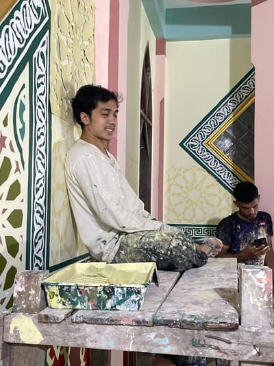 Two men are working on painting a room with intricate geometric designs. One man lies on his back on a patterned floor, his face visible, dressed in paint-splattered overalls. Another man, wearing a paint-covered shirt and sandals, stands atop a scaffold-like structure made of paint-stained boards and containers. He looks at a cell phone. The walls behind them are adorned with similar geometric patterns, and a window is framed by a golden border.