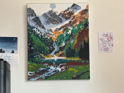 The image shows a wall with several items displayed on it. The central focus is a painting of a mountain landscape with a waterfall and a lake. The mountains are depicted with snow-capped peaks and rocky slopes, with patches of green trees in the foreground. A stream flows from the lake down into the lower part of the painting. To the left of the painting is a smaller framed artwork depicting a night sky with a moon and stars, and silhouetted evergreen trees in the foreground. Below this artwork, a small piece of paper is attached, with handwritten text that appears to be in Russian. To the right of the central painting, a small card with a floral design is attached to the wall. The card features pink cherry blossoms and butterflies, with some text in Russian, including the word "LAQUE" and "WATER STIKER" in English. The overall setting appears to be indoors, likely a room, with the wall painted a light, neutral color. The lighting suggests it is daytime. No people are visible in the image.
