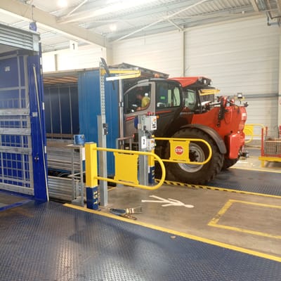 A red and black telehandler is parked in what appears to be a loading dock or warehouse environment. The telehandler has its cab door open, and a person in a yellow shirt is seated in the driver's seat, looking down at a control panel. A yellow safety gate is open in front of the telehandler, and a "STOP" sign is visible on the gate. The floor is a textured blue metal, with yellow safety markings. To the left, a blue loading bay door is open, revealing the interior of a trailer. In the background, industrial shelving and structural beams are visible. A white pedestrian symbol is painted on the floor, and a pair of work straps are lying on the ground near it. The scene is well-lit, suggesting an indoor industrial setting. The location is identified as Ancenis, France.
