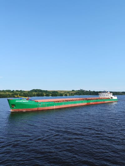 A cargo barge with the name "ДОБРЫНЯ НИКИТИЧ" in Cyrillic letters navigates a wide river under a clear blue sky. The barge is predominantly green with a red waterline. Its cargo hold is covered with what appears to be a rusty-colored tarpaulin. The barge has a white superstructure at its stern.

The scene is set on a calm body of water, likely a river, with a lush green shoreline in the background. Rolling hills covered with trees and some scattered buildings can be seen on the distant bank. The water is a deep blue with subtle ripples.

The bright blue sky suggests it is daytime, likely late morning or afternoon, with fair weather. The overall impression is one of peaceful movement and commerce. The image appears to be taken from a slightly elevated position, looking towards the barge as it travels from left to right. The overall mood is serene and somewhat majestic, highlighting the scale of the barge against the natural landscape.