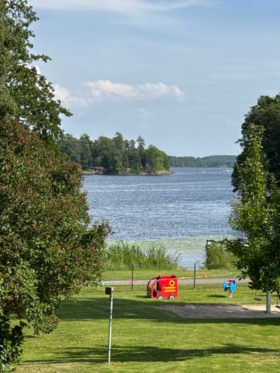 This media file depicts a scenic outdoor setting beside a body of water. The image is oriented vertically, with the water occupying the majority of the left side and a grassy park area to the right. Trees with green foliage frame the top and foreground of the scene.
In the water, there appear to be some faint white objects, possibly sailboats or buoys, and the surface is rippled, suggesting a light breeze. The opposite shore is lined with dense green trees. The park area to the right features a well-maintained lawn and a paved pathway.
On the grass, two toy vehicles are visible. The larger one is a red toy fire truck with the number "112" prominently displayed on its side. A smaller blue toy, possibly a playground structure or another toy vehicle, is located further down the path. A metal pole with a fixture extends from the ground in the grassy area.
The sky above is a pale blue with scattered white clouds, indicating it is daytime. The lighting suggests it is likely a sunny day. The overall impression is one of a peaceful, recreational environment, possibly a park or waterfront area in Askersund, Sweden. There is no indication of people or specific events taking place, but the presence of children's toys suggests it is a family-friendly location. The visible text is "112" on the toy fire truck, which is Sweden's emergency number.