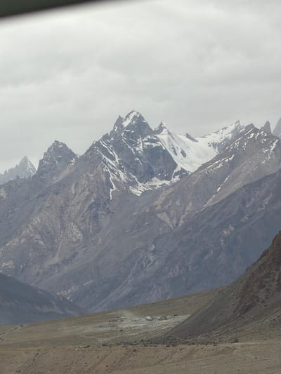 The image depicts a mountainous landscape under a cloudy sky. In the foreground, there is a barren, dusty plain with a winding dirt road. Several small structures, possibly buildings or tents, are visible in the middle ground, with dust plumes suggesting activity or wind. The background is dominated by imposing, rugged mountains with snow-capped peaks. The overall scene conveys a sense of remote, high-altitude terrain. The location is identified as Stongde, India.