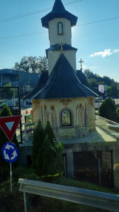A small, Byzantine-style chapel with a dark, tiled roof and a tall, slender tower topped with a spire stands under a clear blue sky. The chapel's walls are adorned with colorful frescoes depicting religious figures, including saints and angels. A narrow, arched window with a mirrored surface is visible on the chapel's facade. Two traffic signs are present: a red and white yield sign and a blue circular sign with white arrows indicating a roundabout. In the background, trees and buildings are visible, including what appears to be a modern commercial structure. The scene is captured from a slightly elevated perspective, suggesting it was taken from a vehicle. The presence of the traffic signs and the commercial building suggests the chapel is located near a road or in a mixed-use area within Fălticeni, Romania.