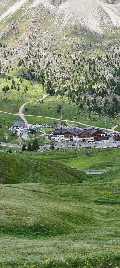 The image shows a panoramic view of a mountain landscape in Selva - Wolkenstein - Selva di Val Gardena, Italy. The scene features a large building complex, likely a hotel or resort, with multiple stories and a dark-tiled roof, situated amidst rolling green hills. Several cars are parked in designated areas around the buildings. In the background, the terrain rises steeply with rocky outcrops and scattered evergreen trees, leading up to a higher mountain peak. Winding dirt paths and ski lift infrastructure are visible on the slopes. The foreground consists of a lush green meadow dotted with small yellow flowers. The sky is not visible in the frame, but the lighting suggests it is daytime.