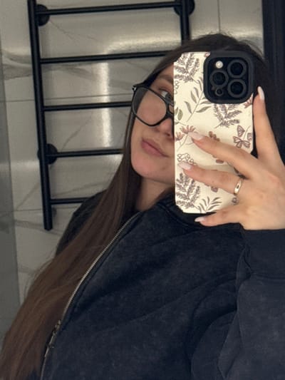 A female individual is depicted taking a mirror selfie indoors. She has long, dark brown hair and wears black-framed eyeglasses. Her attire consists of a dark, possibly faded or acid-washed, zip-up jacket with a visible metal zipper. Her right hand, featuring light-colored nail polish and a plain silver-colored band ring on the ring finger, holds a smartphone. The smartphone is an iPhone model, identifiable by its triple rear camera array, and is housed in a light-colored protective case adorned with a botanical pattern of muted-tone leaves and butterflies. The background, reflected in the mirror, shows light-colored tiled walls and a black, wall-mounted multi-bar towel rack. The precise location within the city of Prikubanskii, Russia, is not ascertainable from the image.