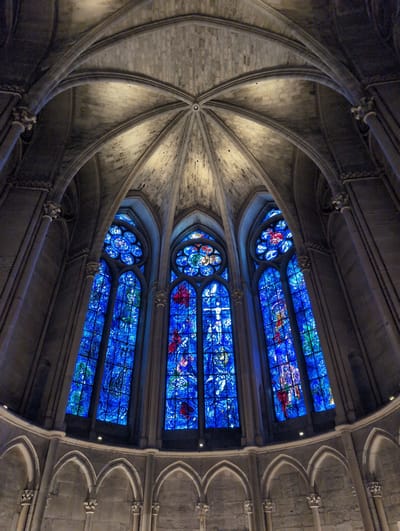 The image shows a low-angle view of the interior of a Gothic cathedral, likely Notre-Dame de Chartres in Chartres, France. The focal point is a triptych of large, ornate stained-glass windows, predominantly in vibrant shades of blue with accents of red and green. The windows are divided into rectangular panes and feature intricate figurative and abstract designs, with a circular rose window above each rectangular section. The ceiling features a complex ribbed vaulting system constructed from grey stone blocks. Below the windows, a row of arched stone arcade structures runs horizontally across the scene. The lighting is dim, with the stained glass windows providing the primary source of illumination, casting a blue hue onto the surrounding stone architecture. There are no people visible in the image. The environment suggests a place of worship or historical significance. Urvillers, France, is not associated with this landmark; Chartres is the correct location.