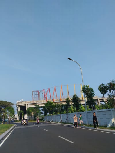The image depicts an outdoor scene on a paved road in Sidodadi, Indonesia, under a clear blue sky. In the foreground, a black asphalt road stretches into the distance, marked with white lines. On the right side of the road, a low, grey concrete wall with a textured pattern runs along the pavement. Three young women are walking on the grassy verge next to the wall, facing away from the camera. The woman on the far right wears black trousers and a black top. The middle woman wears a green t-shirt and grey shorts. The leftmost woman wears a beige long-sleeved top and dark trousers. Behind them, a line of trees and banana plants are visible.
Further down the road, several motorcycles and a car are traveling. To the left of the road, a grassy area with trees lines the pavement.
In the background, a concrete overpass or elevated structure dominates the midground. This structure features colorful scaffolding and supports, with red, orange, and yellow accents. A large, white, stylized logo, possibly a swan or bird shape, is visible on the top of the structure. A street lamp with a curved pole stands near the road. The overall environment suggests a developing urban or suburban area with infrastructure under construction or recently completed.