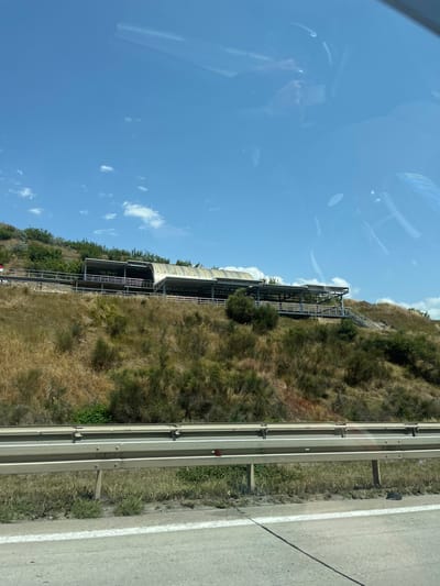 The image depicts a cable car station built into a hillside in Kvemo Rene, Georgia. The station features a long, arched roof structure supported by metal beams and railings. A walkway or platform runs alongside the station, with stairs visible on the right. The hillside is covered in dry, brown vegetation and scattered green bushes and trees. A highway guardrail is in the foreground, separating the road from the hillside. The sky above is blue with scattered white clouds, and there are reflections on what appears to be a car window, suggesting the photo was taken from a moving vehicle.