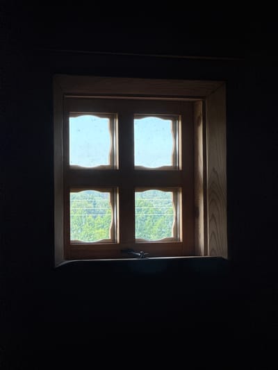 The image shows a wooden-framed window with four panes, set against a dark background. Two panes are visible at the top and two at the bottom, all with a slightly wavy, irregular edge to the glass. The top panes show a bright, hazy sky, while the bottom panes offer a view of green foliage, suggesting an outdoor scene with trees. A dark object, possibly a latch or lock, is visible on the lower part of the window frame. The overall impression is one of a window looking out from a dark interior.
The specific location in Нёманскі сельскі Савет, Belarus, is not discernible from the visual information in the image itself. The image captures a static interior view through a window.