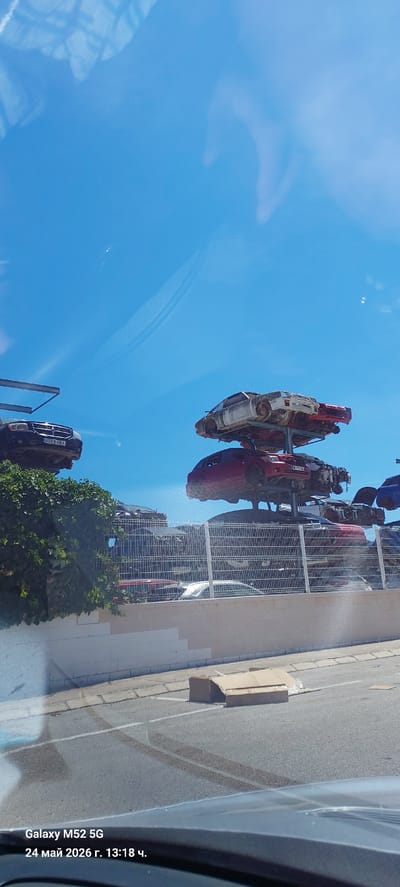 The image shows a car junkyard in Sant Joanet, Spain. The photo was taken from inside a vehicle. The sky is blue and clear. There are several wrecked cars stacked on metal frames. A building is visible behind the car frames, and there is a bush. A paved road is visible on the right side of the image with some cardboard boxes. The image timestamp indicates it was taken with a Galaxy M52 5G on May 24, 2026, at 13:18.
