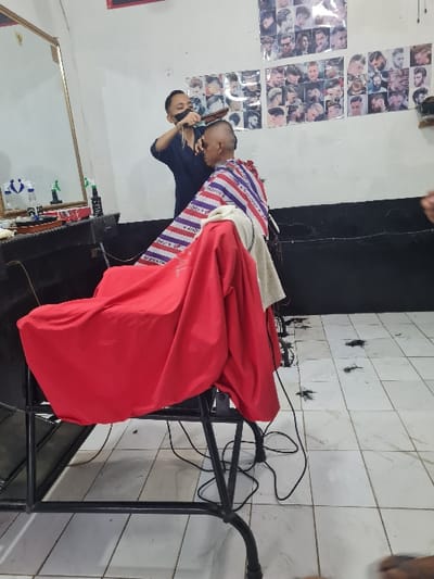 Inside a barbershop, a barber cuts a client's hair with an electric razor. The client is draped in a striped and red cape, seated in a black metal chair. The room features tiled walls with scattered hair clippings, a black wall section, and a mirror. Posters displaying various hairstyles adorn a white wall.