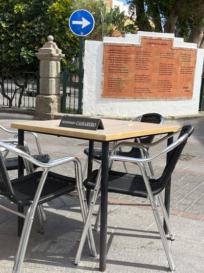 The image shows a table with four chairs in front of a white wall with a plaque on it. The table is made of wood and metal, with a sign on it that says "Reservado Camarero." The chairs are silver metal with black woven seat and backs. The plaque is made of brown ceramic tiles with white writing. It depicts a poem in Spanish titled "El Romance de Agüimes."  The scene is located at 27.907, -15.447 in Agüimes, Spain. The environment is a public square, with a cobblestone pavement and green plants in the background. There are no people in the image.  The image is taken from a perspective that looks down onto the table and chairs. The chairs are empty, suggesting that the table is reserved for a waiter.  The writing on the plaque is visible but not legible from the image.