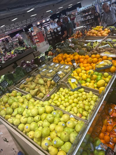 This image depicts a produce aisle in a supermarket, likely located in Ardea, Italy, given the presence of Italian signage like "Gastronomia" and prices in Euros. The scene is filled with various fruits and vegetables displayed in wooden crates and clear plastic packaging. Prominently featured are large quantities of pears, oranges, lemons, limes, bananas, pineapples, kiwis, red cabbages, and eggplants. Several people are visible browsing the produce and shopping. On the right side of the image, a man wearing glasses and a blue checkered shirt is looking at the produce. To his left, a man in a dark shirt and a younger man in a black t-shirt are also present. Further back, a woman with dark hair is seen near a display. The environment appears to be well-lit with overhead fluorescent lighting. The overall impression is a busy, well-stocked grocery store section.
