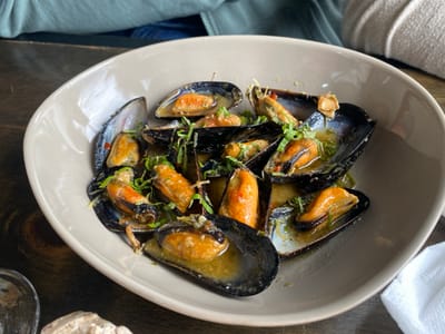 The image displays a close-up view of a meal served on a dark brown wooden table, consistent with an indoor dining establishment in Puerto Almanza, Argentina. The primary subject is a large, light beige-colored bowl containing approximately 15 to 20 cooked mussels. Each mussel is presented in its dark blue-black shell, with most shells open to reveal the orange-hued mollusk meat. The mussels are partially submerged in a clear, yellowish-green broth or sauce and are garnished with finely chopped green herbs, likely parsley, along with small white fragments that may be garlic. In the upper left quadrant, the shoulder and arm of an individual wearing a teal-colored long-sleeved garment are visible, indicating a person seated at the table. A clear glass is partially visible in the lower left foreground, next to an irregularly shaped, light-colored object, possibly a piece of bread. A folded white napkin or cloth is partially visible in the lower right background. No explicit actions or interactions are depicted beyond the implied act of dining.