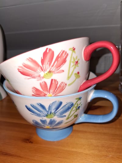 Two ceramic mugs are stacked on a wooden surface. The top mug is white with red floral designs and a bright pink handle. The bottom mug is also white with blue floral designs and a light blue handle. The mugs appear to be in a home setting, possibly on a table or shelf. The image does not provide enough information to determine the specific location within Åsgårdstrand, Norway, or any details about people, actions, or interactions.