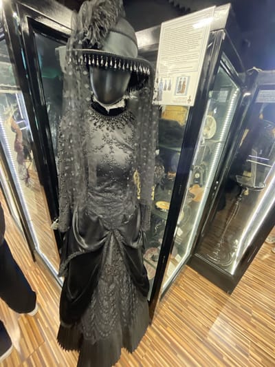 In the city of Voskhod, Russia, inside what appears to be a museum exhibit, a black mannequin is displayed wearing a formal black gown and a matching hat. The dress is ornate, featuring intricate beading on the bodice and sleeves, a satin skirt with draped detailing, and a lace hem. The hat is adorned with feathers and beaded trim, and a veil partially obscures the mannequin's head. The mannequin stands in front of a glass display case that contains various artifacts, including framed images and small decorative items, illuminated by bright lights. To the left and right of the main display, other glass cabinets are visible, also containing exhibits. The floor is made of polished wood with a striped pattern. A person's legs, clad in dark pants and white sneakers, are partially visible in the lower left corner of the frame, suggesting a visitor to the exhibit.