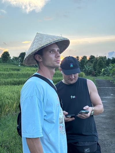 Two Caucasian men stand close to each other in Padangbulia, Indonesia. The man in the foreground wears a light blue t-shirt and a traditional conical hat. The man behind him wears a black sleeveless shirt with "fw." printed on it, as well as a black baseball cap. He's holding two black, rectangular objects, possibly cell phones, in his hands. The background consists of a field of tall grass, trees, and a path, with a sunset visible in the sky.
