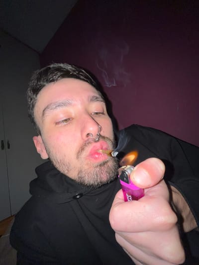 The image shows a man with fair skin, light-colored eyes, dark hair, and a beard, holding a lit cigarette in his mouth. He is wearing a black hoodie. He has a septum piercing. In his right hand, he is holding a pink lighter with a flame. Smoke is visible emanating from the cigarette. The background appears to be a wall with two different colors: dark purple at the top and white at the bottom. The location of the scene is not discernible within Llavallol, Argentina.