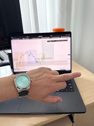 A person's wrist wearing a silver watch with a turquoise dial is in the foreground, resting on a laptop. The laptop screen displays a financial chart with various colored blocks and lines. The laptop is on a light-colored wooden desk. In the background, white vertical blinds are visible, and a portion of a dark object, possibly a monitor, is to the left. An orange circular object is positioned on top of the laptop, near the screen. The scene appears to be indoors, likely a home office or workspace, though no specific location in Vaughan, Canada, can be identified from the image alone.