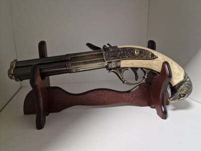 A decorative double-barreled pistol with an ornate, ivory-colored grip carved in the shape of a wild boar's head is displayed on a wooden stand. The pistol has a dark metal finish with intricate metalwork on the barrel and frame. The stand is made of polished dark wood and has two upright supports to hold the pistol. The pistol and stand are placed on a white shelf, which is part of a white shelving unit against a plain white background. The lighting is even, casting soft shadows. There are no people or any other objects visible in the image. The scene is indoors, likely in a home or a display setting. The location of Pogranichny, Russia is not determinable from the visual information provided.
