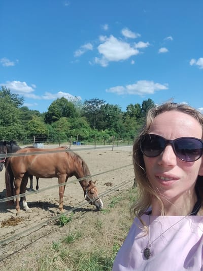 The image is a selfie taken outdoors with two horses in the background. The person in the foreground is wearing sunglasses and a light purple shirt with a silver pendant necklace. The background shows a sandy paddock with a fence and several trees. The sky is blue with some white clouds. The scene appears to be a farm or equestrian center. The location of Winterswijk Huppel, Netherlands is not explicitly visible in the image but is provided as context.