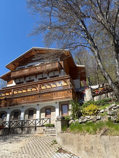 The image shows a three-story building with wooden balconies, likely a guesthouse or hotel named "Koprivshtica," situated in Ichera, Bulgaria. The facade is a combination of white and stone with arched windows on the ground floor. Wooden railings are present. A tree with bare branches occupies the upper left corner of the image, contrasting with the clear blue sky. A stone wall with vegetation runs along the right side of the building, adjacent to a paved area constructed of square blocks with holes in the center. An additional structure is visible behind the main building.