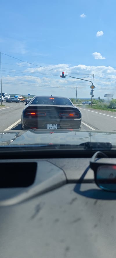 A rear view of a dark-colored Dodge Challenger with the license plate "T 173 XY 73" is visible through the windshield of another car. The Challenger is stopped at a traffic light in Prigorodny, Russia. The traffic light is red. The sky is mostly clear with scattered clouds. Other vehicles are visible in the background. Road signs are present on the right side of the road. Sunglasses are on the dashboard of the car taking the photo.