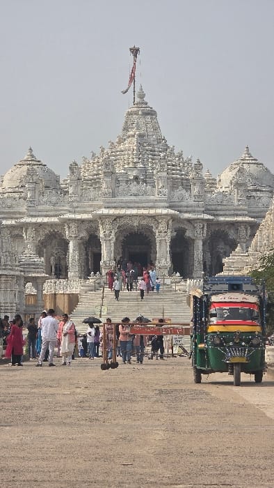 A large, ornate white temple rises on the left, its spires and intricate carvings prominent. People gather near the entrance, ascending the steps. A colorful auto-rickshaw is parked on the right side of the temple. A signpost in front of the steps indicates directions in a language other than English. The temple and auto-rickshaw are situated beside a long paved path. The photograph is rotated counterclockwise, with the path running vertically.