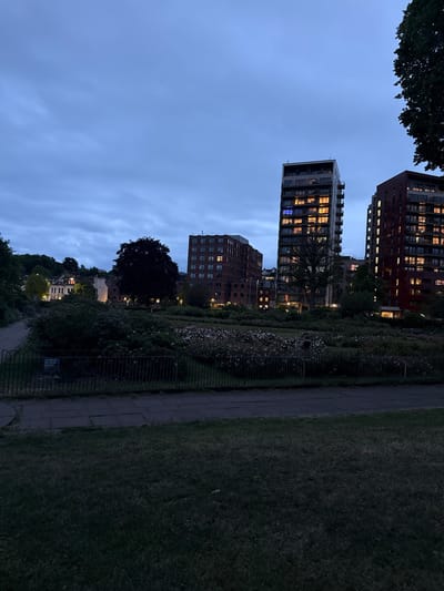 The image depicts a park in Preston, United Kingdom, at dusk. Three high-rise buildings with illuminated windows are visible. A tree and greenery can be seen along a walkway. The sky is overcast.