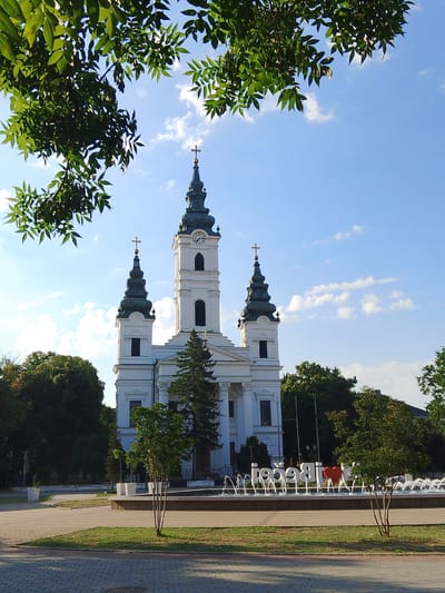 In the city of Bečej, Serbia, a white church with multiple spires, one of which has a clock, stands tall against a bright blue sky with scattered clouds. Lush green trees frame the top of the image, and smaller trees are visible in front of the church. In the foreground, a fountain with several jets of water is active. Beyond the fountain, a large sign with the words "I love Bečej" is displayed, featuring a red heart. The ground is paved with stone in the foreground and has grass and paving stones in the middle ground. The scene appears to be a public square or park.