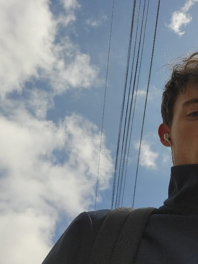 The image shows a young man standing with his back to the camera, looking up at the sky. He is wearing a black turtleneck sweater and a black backpack. The sky is blue with white clouds. The photo was taken on a street in Calais, France, at the coordinates 50.955 latitude and 1.859 longitude. There are multiple power lines running across the image diagonally from the top left corner to the bottom right corner.  The young man has a white earbud in his right ear.