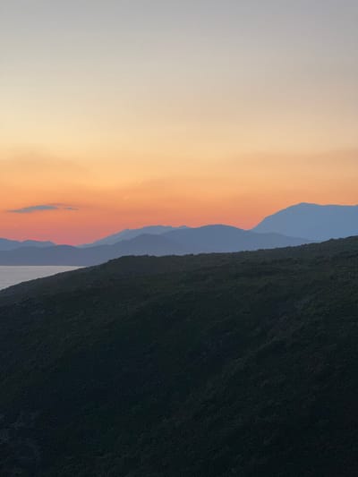 The image depicts a serene sunset over a mountainous coastal landscape, likely in Glavatičići, Montenegro. The sky transitions from a soft pale blue at the top to a warm gradient of orange and pink hues near the horizon. Silhouetted against the colorful sky are several layers of mountains, receding into the distance. In the foreground, a dark, densely vegetated hillside slopes downwards towards a body of water, possibly the Adriatic Sea, which is visible on the left side of the frame. The overall scene is tranquil and natural, characterized by its natural beauty and subdued lighting. There are no people or man-made objects visible in the frame, emphasizing the unspoiled, natural environment.