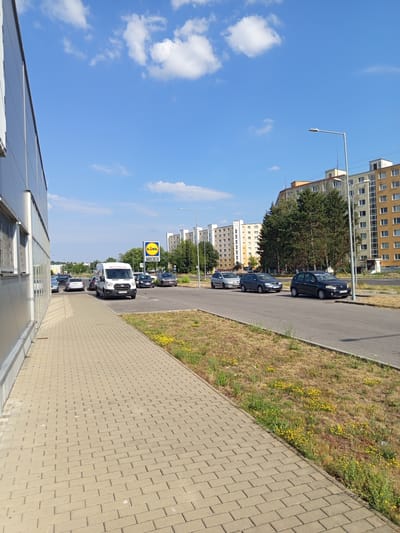 This is an outdoor, eye-level shot of a street in Zvolen, Slovakia, on a sunny day. The camera is tilted slightly, looking down the street which is lined with buildings on one side and a grassy embankment with yellow flowers on the other. Parked cars and a white van are visible along the roadside. A bright yellow Lidl sign is also visible. The sky is a clear blue with a few scattered white clouds. The buildings appear to be apartment complexes. The foreground shows the side of a modern building with reflective windows.