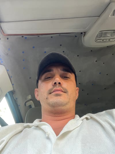 The image shows a medium shot of a man wearing a baseball cap and a white collared shirt. He is looking directly at the camera. The man has short dark hair, a mustache, and a light beard. He is wearing a silver earring in his left ear. The background appears to be the interior of a vehicle, specifically the headliner, which is covered in a gray fabric with numerous small blue dots. There is also a dome light visible on the headliner. The lighting suggests it is daytime. The image quality is decent, with good focus on the man's face. The scene is static, with no clear action or interaction depicted. The location is not specified in the image itself, nor can it be inferred from any visual cues within the frame. Therefore, it cannot be definitively stated that this image was taken in Gornja Brova, Montenegro.