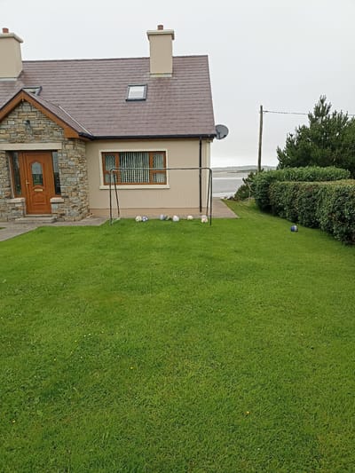 This is a medium-range exterior shot of a suburban house in Annagry West, Ireland, on an overcast day. The house, a two-story structure with a stone facade on the front left and beige siding on the rest of the exterior, features a brown tiled roof with two chimneys and two skylights. A wooden door with glass panels is visible on the front left.

In front of the house, a well-maintained green lawn stretches out, dotted with small white and yellow flowers. A small soccer goal with a net is set up on the lawn. Several soccer balls are scattered around the goal, including one blue and white ball on the far right of the lawn. Behind the house, a dense green hedge lines the property, and beyond that, a body of water is visible, likely the Atlantic Ocean, with a distant shoreline under a gray sky. A single utility pole with wires stands to the right of the house.