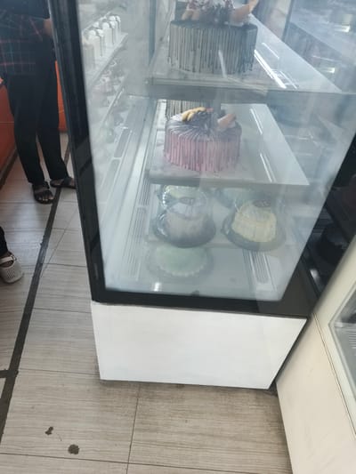 In Krian, Indonesia, a bakery display case showcases a variety of cakes. The case is lit from within, highlighting the desserts. On the top shelf, a chocolate-drizzled cake with floral decorations sits prominently. Below it, a pink striped cake adorned with fruit and chocolate pieces is visible. Further down, several smaller cakes are displayed in individual clear plastic containers. Two individuals are partially visible near the display case. One person, wearing a plaid shirt and dark pants, stands to the left, their feet in sandals visible on the tiled floor. Another person's feet, wearing white sandals, are also visible on the floor to the left of the frame. The tiled floor has some dark stains. The display case has a white base unit.