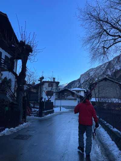 This image captures a bright, clear winter day in Courmayeur, Italy, depicting a snowy alpine village scene. The sky is a vibrant, cloudless blue.

The primary subject is a person, seen from behind, walking on a wet, paved road or path. They are wearing a bright red jacket, grey pants, and dark shoes, and are carrying a pair of skis and ski poles over their right shoulder. The road shows patches of snow and slush along its edges.

The setting features traditional chalet-style buildings with stone and wooden elements, some having balconies. These structures line both sides of the path. In the foreground, on the left, a large, gnarled tree with bare, dark branches dominates the lower left corner and extends into the sky. Other bare trees are scattered throughout the scene.

In the midground, a two-word wooden sign hangs from one of the buildings; the text appears to read "Snappy Ray". Beyond the buildings, majestic, snow-dusted mountains rise in the distance under the clear blue sky. The overall impression is of a crisp, sunny winter day, suggesting conditions favorable for skiing.