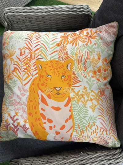 A close-up, high-angle shot shows a decorative cushion with a leopard pattern on a dark gray outdoor sofa. The cushion is predominantly white with an orange and green floral and leaf print. In the center of the cushion is a stylized orange leopard's head and chest, with orange spots. The leopard has green eyes and whiskers. Behind the leopard, the background is filled with various tropical-looking leaves and flowers in shades of orange, green, and pink. The cushion has a thin green trim along its edge. To the left of the cushion, a portion of a gray rattan chair is visible. The background is slightly blurred, suggesting an outdoor setting, possibly a garden or patio. No people are present in the image. The scene is static and indoors or outdoors with artificial light. The image does not depict any specific landmarks or context that would allow for identification of a city in Belfast, United Kingdom.