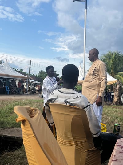 An outdoor community gathering is captured in a portrait-oriented shot, featuring several individuals and event infrastructure under bright daylight. In the immediate foreground, a person's back is visible, draped in a flowing gold-colored garment. Adjacent, another individual is seated, seen from behind, wearing a grey hoodie adorned with black and white geometric patterns, and the text "machout bothcs" and "1st" on the back.

Further into the mid-ground, a person wearing a white long-sleeved shirt and a black cap is actively speaking into a microphone. To their left, a man in a tan/light brown traditional garment over white trousers stands, observing the event. The background reveals more attendees seated on blue plastic chairs under white tents.

The setting appears to be a rural or semi-urban area, characterized by a dirt and grass ground, lush palm trees, and other green foliage. Utility poles with power lines run along the left side, and a simple building complex with parked motorcycles and a white truck is visible in the distance. The sky is bright blue with scattered white clouds, indicating clear weather. A green "33 LAGER" beer can and a yellow plastic item are on the ground near the seated figures. The overall scene suggests a public address or social event taking place in Aghalokpe, Nigeria.
