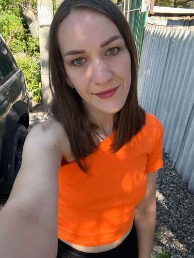 A woman with long brown hair and green eyes, wearing an orange t-shirt and black pants, takes a selfie outdoors. She is standing next to a dark-colored SUV, with a corrugated metal fence and gravel on the right side of the frame. Lush green foliage and a tree are visible in the background. The scene appears to be in a residential or rural area, possibly in Vinsady, Russia.