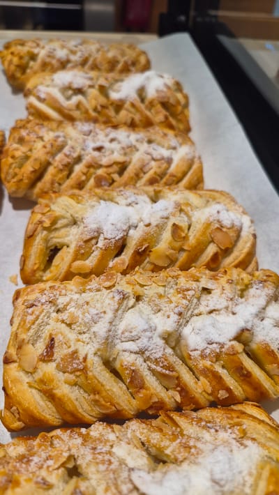 A close-up, vertical shot shows a row of golden-brown, braided pastries dusted with powdered sugar and topped with sliced almonds. The pastries are arranged on a white surface, likely parchment paper, which is resting on a dark, reflective surface. The lighting is warm and highlights the flaky texture of the pastry and the sheen of the almonds. The background is blurred, but hints at a commercial setting with dark vertical elements, possibly shelving or a counter. The pastries appear to be a sweet baked good, possibly a type of danish or strudel, prepared for sale or display. The image does not provide any information about the location in the city of Līvāni, Latvia.