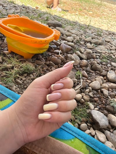 In Koktobe, Kazakhstan, a close-up shot captures a hand with light skin and meticulously manicured fingernails adorned with a French tip style, featuring a pale yellow hue on the tips and a sheer pink base. The hand rests on a striped fabric in shades of blue and green. The background is a natural setting comprised of river stones and sparse green vegetation. An orange and yellow plastic toy bucket, containing murky water, sits amongst the stones, suggesting a playful or leisurely activity. The overall scene is outdoors, under daylight conditions, and implies a casual, recreational environment near a body of water.