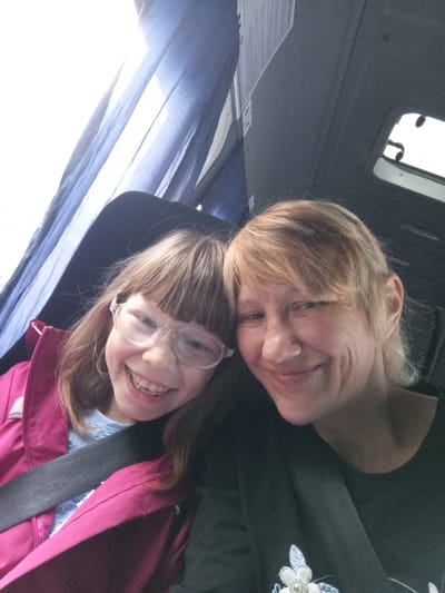 Two individuals, a child and an adult, are positioned in what appears to be the interior of a vehicle, possibly a bus or van, as suggested by the presence of seatbelts and interior paneling. The child, on the left, is wearing a bright pink jacket over a light blue shirt and has on clear-rimmed glasses. They are smiling broadly, revealing braces on their teeth. The adult, on the right, has blonde hair and is wearing a dark shirt with a floral design. Both are looking towards the camera, indicating they are taking a selfie. The background shows blue curtains and grey interior surfaces. The scene suggests a casual moment of travel. The location within Verkhneyarkeyevo, Russia, is not discernible from the visual information provided in the image.