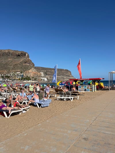 The image shows a beach in Los Cristianos, Tenerife, Spain. The beach is crowded with people, many of whom are lying down on beach chairs or walking around. There is a red-and-white striped awning with a sign that reads "www.aquasports.es". There are also several colorful inflatables in front of the awning. The background of the image features a large cliff with a small town nestled at its base. The sky is blue with no clouds.